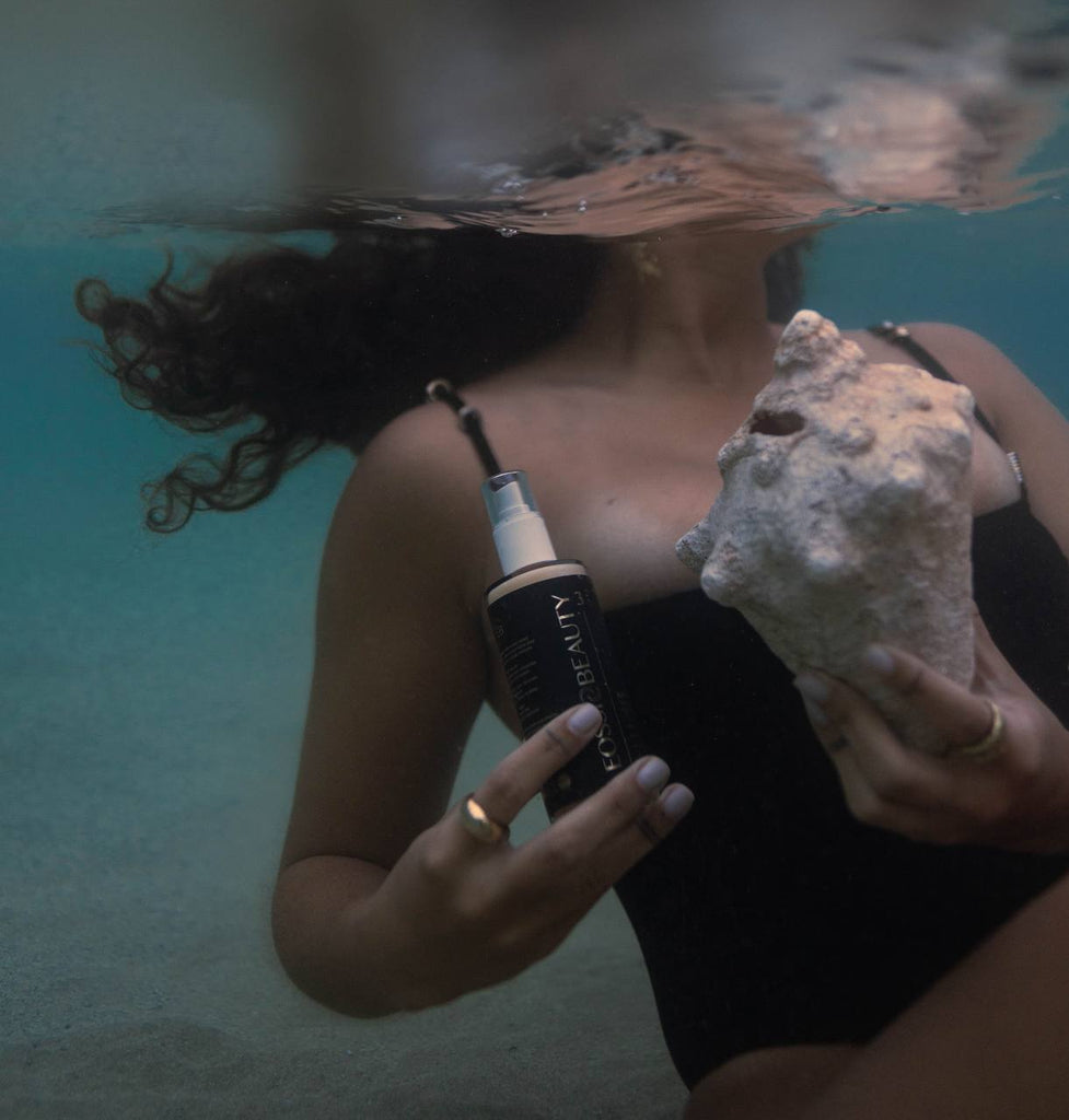 Woman in a black bathing suit holding a conk shell and a glass bottle with a black and gold label on it submerged in the ocean.
