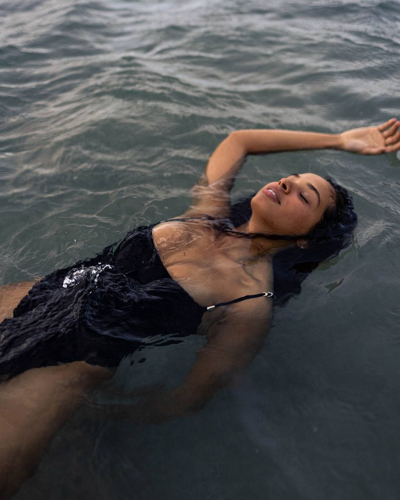 woman floating in the ocean in a black bathing suit with her arm up and her eyes closed