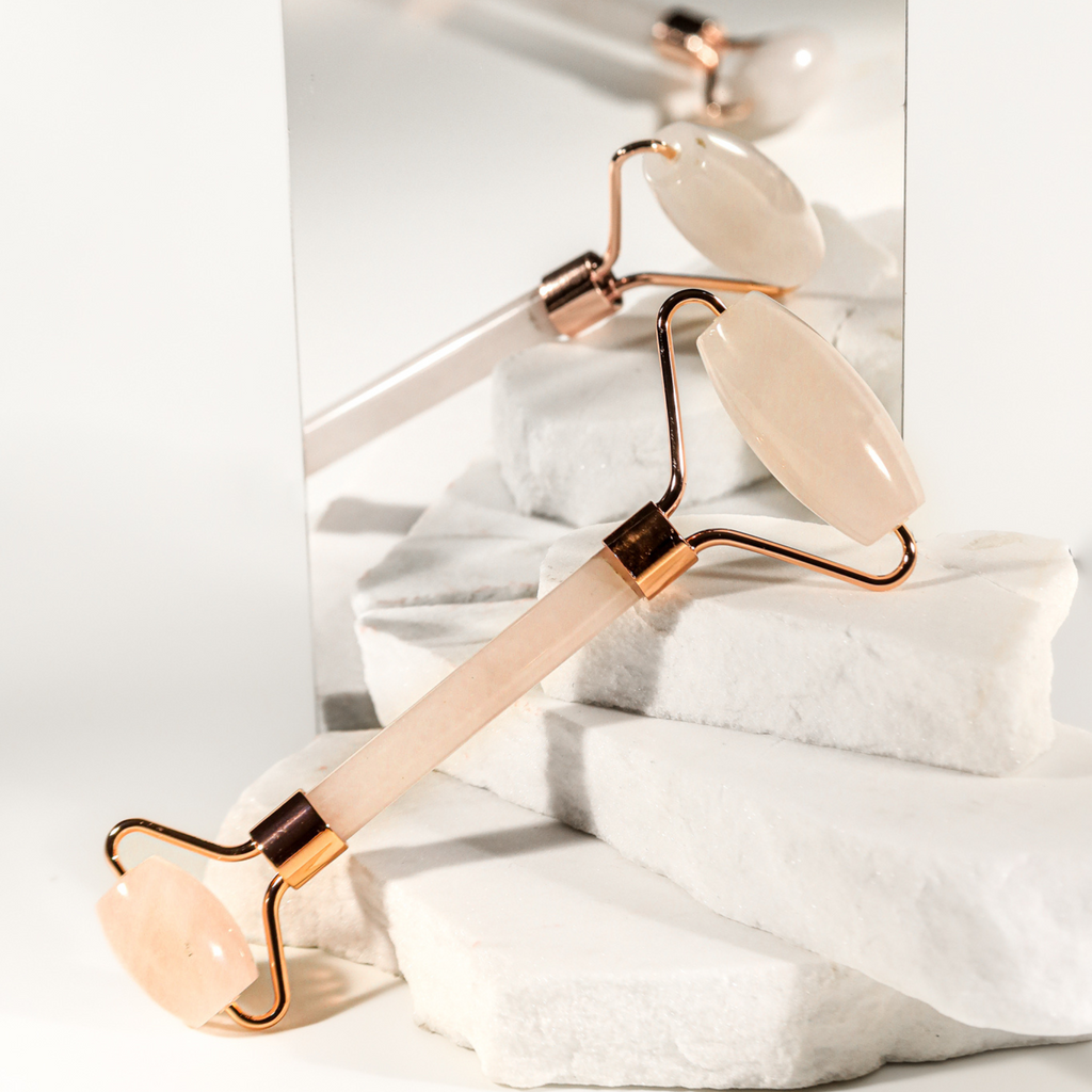 Rose quartz facial roller with gold hardware, resting on marble stairs against a mirrored background, for skincare routine.