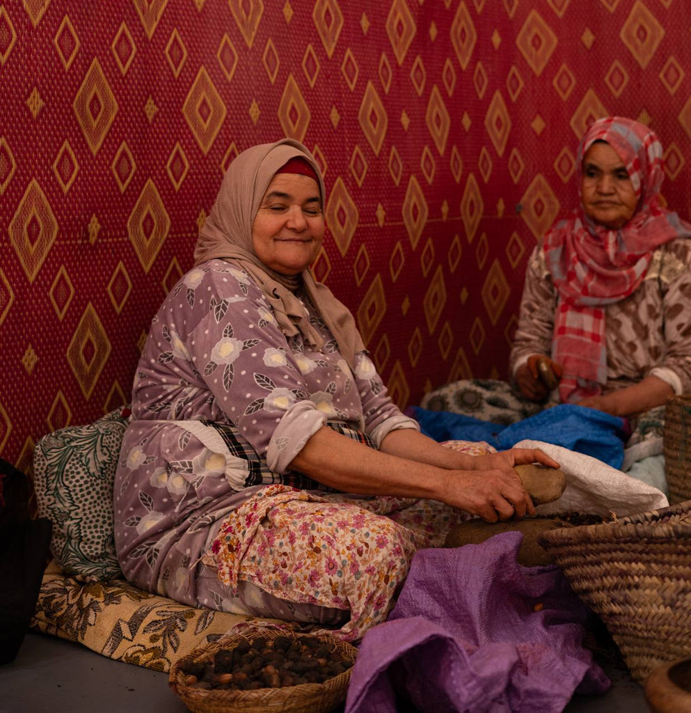 Two Moroccan women in traditional attire handcrafting Argan oil in a cooperative, surrounded by baskets of Argan nuts, showcasing their skill and cultural heritage