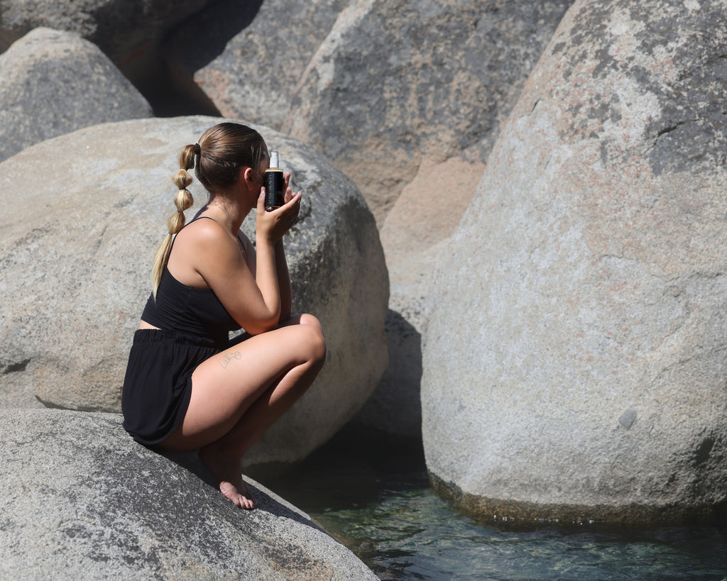 A woman in a black shorts and a black tank top perched on a large, smooth boulder by the water, examining a glass bottle with product from the Eossi Beauty. She's captured in a moment of focus, with a natural backdrop of rounded stones and a calm water surface, suggesting an environment of a pure and mindful skincare routine.