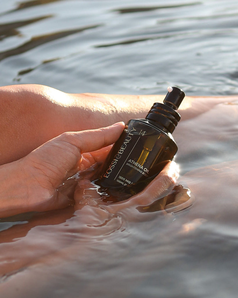 Hand gently holding Eossi Beauty body oil bottle partially submerged in tranquil water, symbolizing the product's hydrating properties.