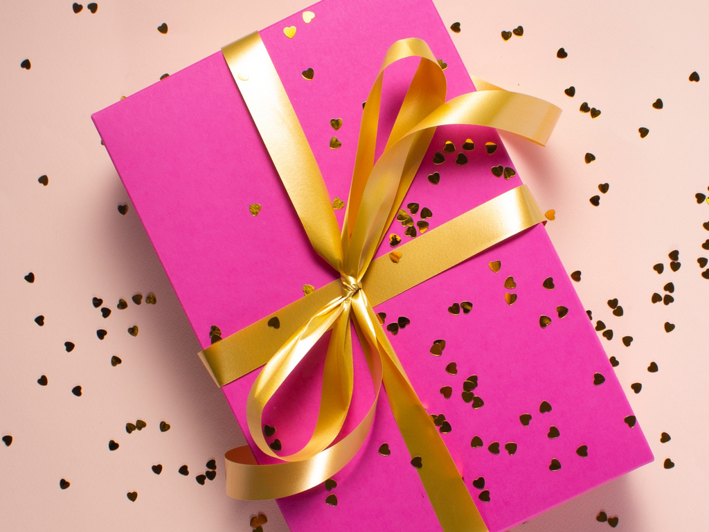Dark pink gift card box with a gold ribbon around the box on a light pink background with gold heart shaped confetti strewn around the image.
